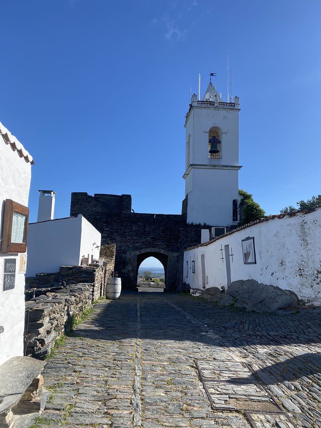 The Porta da Vila seen from inside the village. The original bell, dating from 1692, is housed in the tower on the right.|There are only two streets in Monsaraz that run the length of the village. This is the main one. Wheeled vehicles are not allowed, except for loading/unloading, but there is plenty of parking just outside the walls.|Another view of the Rua Direita. All the village’s streets and lanes are cobbled.|This ATM, next to the Junta da Freguesia, is about the only concession Monsaraz makes to the 21st century.|The main tower of the castle, called the Torre de São Gens do Xarez, or, more familiarly, Torre das Feiticeiras (Tower of Witches). Construction of the castle was started by King Alfonso III and eventually completed by his son Dinis I in the early 13th century. Entrance is free. The views from the tower are exceptional.|One of the many beautiful views from the village – this one looking east to the Alqueva Lake.|A reminder that the Templar knights were active in the castle’s construction and defense.|The fertile Alentejano plains as seen from the castle|Another view of the village. Note the absence of tourists and of cars!|The Porta da Cisterna and, beyond it, the Porta do Buraco, one of the four entrances to the village.|The hilltop walled village of Monsaraz, seen from the east (the Alqueva Lake) side. The castle and the Igreja Matriz are clearly visible, anchoring the walls.|The 16th century Igreja Matriz, Nossa Senhora da Lagoa, which was rebuilt after the 1755 earthquake and is currently being renovated. In front of the church, in the main square (Largo Nuno Álvares Pereira), is an 18th century marble pillory (pelourinho) where announcements were made and laws and punishments given.|A view over the roofs of Monsaraz, looking southeast towards the Alqueva.|Helga and Robbie Edwards toast Monsaraz with a lovely local vinho branco on the terrace of the Sahida restaurant.|The Porta da Vila serves as the main entrance to the walled village. It dates from the 17th century. Above the arch, there is a memorial to the Immaculate Conception, installed by King João IV in 1646