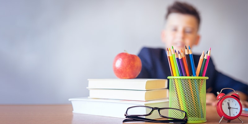 Blurred image of a boy sitting at a desk with books, pencils and a pair of glasses to symbolise the importance of doing an eye check-up in children||View of the outside of Optimax's store in downtown Faro