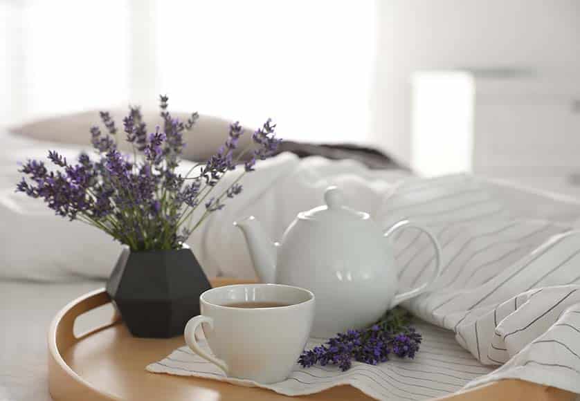 Wooden tray with cup of hot tea and beautiful lavender flowers on bed. Tasty breakfast