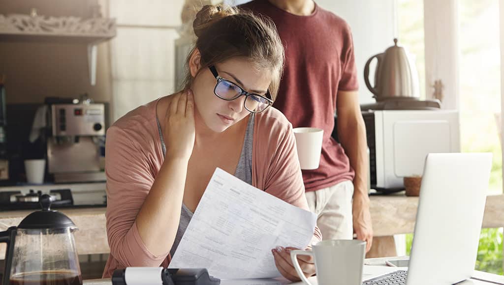 Unhappy beautiful woman wearing spectacles having concentrated look reading notification form bank on debt, sitting at kitchen table in front of open laptop, her husband drinking coffee on background|Electricity – understanding your bill