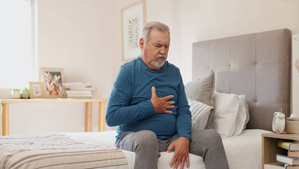 Senior man, chest pain and injury with heart condition or panic attack in bed at home. Elderly male person with ache, sore body or cardiac arrest in stress, anxiety or health in bedroom at house