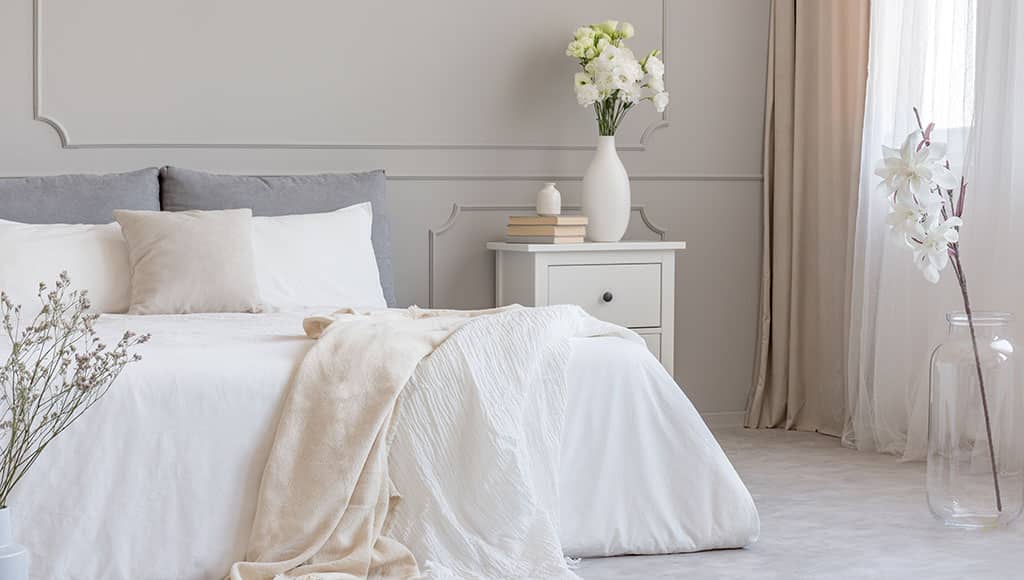 Roses in white stylish vase and pile of books on wooden nightstand with drawers next to cozy bed with blanket
