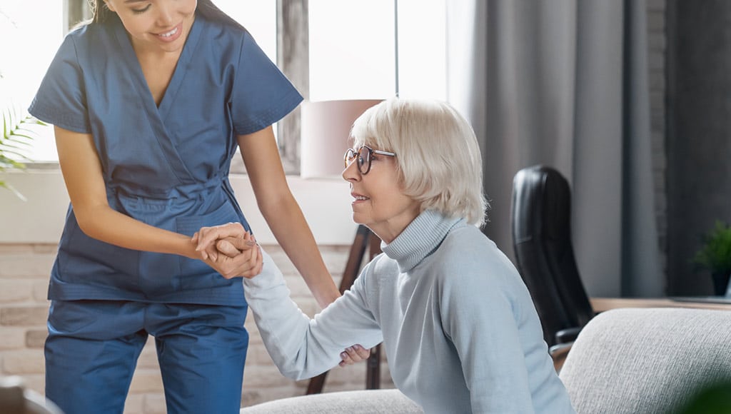 A young woman providing live-in care and helping an old home get up from her sofa