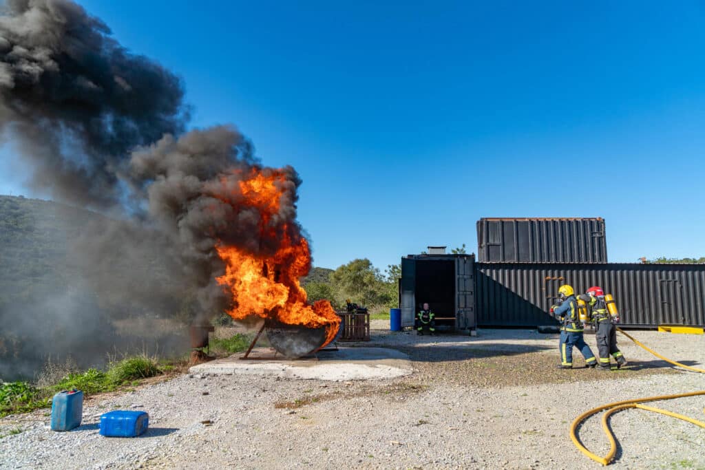 Bombeiros trainees approach fuel fire bursting with flames and spewing thick black smoke